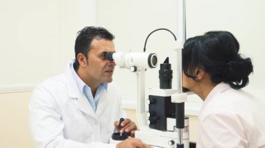 Doktor, sıcak bir gülümsemeyle, hastaların gözlerini incelikle inceliyor sofistike bir görüş testi cihazıyla. Hasta, genç bir kadın, rahatça başını aparatta dinlendiriyor, göz doktorlarında güven verici bir atmosfer yaratıyor.