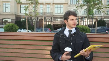 Şık siyah takım elbiseli genç bir adam ahşap bir bankta oturuyor, parlak sarı klasöründeki belgeleri dikkatle gözden geçiriyor. Sıcak bir tebessümle, kahve fincanından bir yudum alır. Etrafı yemyeşil ve zarif bir park sahnesiyle çevrilidir.