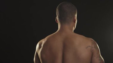 Güçlü bir Afrikalı adam dikiz aynasıyla kendine güvenerek ısınıyor, etkileyici sırt kaslarını omzunun üstünden geçerken esnetiyor. Dramatik ışıklandırma onun tonlu fiziğini vurguluyor, bir spor salonundaki güç ve kararlılığını gösteriyor.