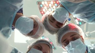 Tıbbi maske ve şapkalar takmış yetenekli bir cerrahi ekip hastalarının üzerine eğiliyor ve uzmanlıklarını ve takım çalışmalarını sergiliyor. Bu yakın plan bakış açısı, dört erkek cerrah ve bir kadın hemşirenin yüzündeki kararlılığı yansıtıyor.