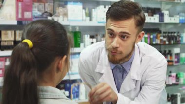 Canlı ürünlerle dolu parlak bir eczanede neşeli bir eczacı güzel paketlenmiş ürünleri küçük mutlu bir kıza veriyor. Dost canlısı bir adamla kızın, annesinin yanında sıcak bir etkileşim oluşturması çok iç açıcı.