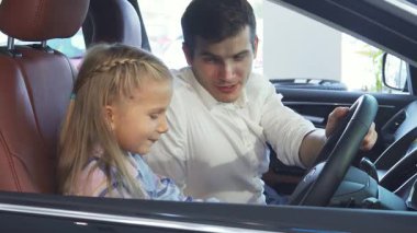 Neşeli bir baba ve küçük kızı şık bir arabanın içinde oturup hoş bir sohbete dalarlar. Sıcak güneş ışığı pencerelerden süzülüyor, gülümsemelerini aydınlatıyor ve sevgi ve bağlantı dolu samimi bir atmosfer yaratıyor.