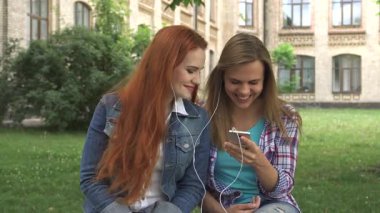 İki neşeli kız öğrenci, biri canlı kızıl saçlı, diğeri açık kahverengi saçlı, çimenli bir kampüs çimenliğinde oturuyorlar, akıllı telefon ve kulaklıkları paylaşıyorlar. Kahkahaları, yemyeşil yemyeşil, güneşli bir günün tadını çıkarırken havayı dolduruyor.