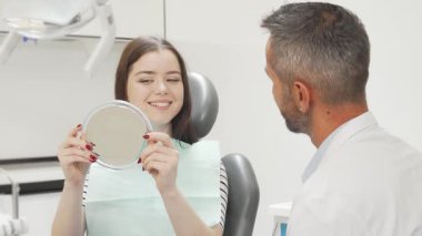 Neşeli bir kadın, aynada ışıl ışıl gülüşünü hayranlıkla seyrederken dişçisiyle dostane bir sohbete dalıyor. Diş kliniğindeki bu canlandırma sahnesi dişçi muayenesinin başarılı sonuçlarını ön plana çıkarıyor.