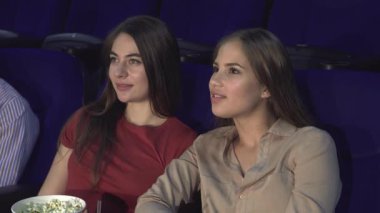 Samimi bir sinemada yan yana oturan iki genç kadın, neşeli bir fısıltı ve gülümsemeyi paylaşırken ekranda parlayan yüzleri. Patlamış mısır ve heyecan havadayken, bu keyifli an arkadaşlığın ve