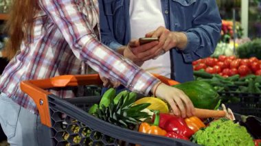 Genç bir çift hareketli süpermarkette canlı sebzeleri ve meyveleri coşkuyla ayırıyor. Kadın olgun bir domates tutarken, erkek arkadaşı da akıllı telefonuna sağlıklı bir şey kaçırmamalarını sağlamak için bir şeyler not alıyor.