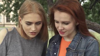 Biri çarpıcı kızıl saçlı, diğeri sarı bukleli iki genç kadın üniversitelerinin yakınındaki yemyeşil bir parkta mutlu bir şekilde akıllı bir telefonu paylaşıyorlar. Birlikte interneti keşfederken kahkahaları havayı dolduruyor, canlı bir atmosfer yaratıyor.