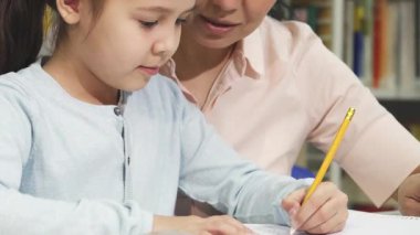 Yürek ısıtan bir yakın plan, ev ödevleriyle yakından ilgilenen neşeli küçük bir kızı yakalar ve destekleyici annesi yanında sevgiyle gülümserken. Bu samimi an, sıcak bir ev ortamında öğrenmenin ve kaynaşmanın güzelliğini gösteriyor.