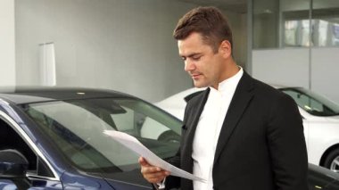 Yeni arabasının anahtarlarını elinde tutan bakımlı sakallı keyifli bir adam modern bir galeride lüks araçlarla çevrili. Gıcır gıcır siyah takım elbisesi ve kareli gömleği bu önemli alışverişe duyduğu heyecan ve güveni yansıtıyor.