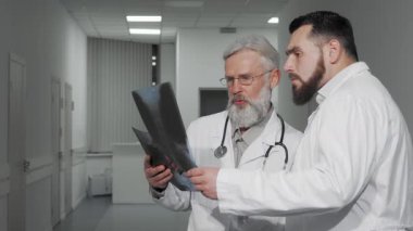 Gösterişli, iyi aydınlatılmış bir klinik koridorunda, iki erkek doktor hastanın röntgen taramalarını incelerken odaklanmış bir tartışmaya giriyorlar. Kıdemli pratisyen, seçkin sakallı ve gözlüklü, genç asistanıyla işbirliği yapıyor,