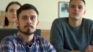 Yakışıklı, sakallı bir erkek öğrenci, derinden odaklanmış, modern bir konferans salonunda zarif bir dijital tabletle etkileşime geçiyor. Kamera konsantrasyonunun yoğunluğunu yakalıyor, ondan en ön sırada güzel bir kız öğrenciye geçiyor,