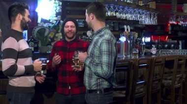 Üç çekici beyaz adam canlı bir barda neşeli bir kadeh kaldırarak kadehlerini kaldırıyor. Etrafı canlı bir atmosferle çevrili. Şapkalı yakışıklı sakallı adam arkadaşlarıyla gülüyor, dostluk dolu sıcak ve davetkar bir sahne yaratıyor.