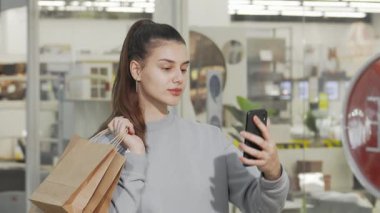 Neşeli genç bir kadın akıllı telefonuyla selfie çekiyor, şık alışveriş merkezlerinde şık alışveriş torbaları tutuyor. Bu canlı sahne mutluluğu yansıtıyor ve modern tüketiciliğin özünü yansıtıyor.