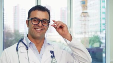 Genç doktor, sıcak bir gülümsemeyle, parlak ofisinde kendine güven ve yakınlık yayarak gözlüğünü çıkarıyor. Doğal ışık ve modern dekor ile çevrili, hastalarına yardım etmeye hazır, sağlık ve bakımı somutlaştırıyor.