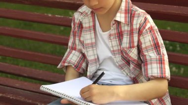 Ekose gömlek giymiş sevimli küçük bir çocuk canlı yeşil bir çimenlikte defterine çizim yapmaya derinden odaklanmış. Güneş ışığı etrafında dans ediyor, açık havada yaratıcılığın keyifli anlarından zevk alırken neşeli yüzünü aydınlatıyor.