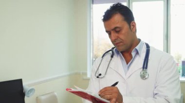 Genç erkek doktor, iyi aydınlatılmış ofisinde güven içinde duruyor ve defterine önemli notlar yazıyor. Boynunda steteskop ve sıcacık bir gülümsemeyle hastalarına yardım etmeye hazır profesyonellik ve ilgiyi somutlaştırıyor.