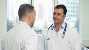 Birinde steteskop, diğerinde beyaz önlük olan iki doktor sıcak bir el sıkışma, tatmin ve dostluk paylaşıyor. Gülüşleri, parlak ve modern ofis ortamında işbirliği üzerine başarılı bir tartışmayı yansıtıyor.