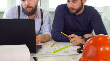 Birinin esmer sakalı ve pantolon askısı olan iki genç erkek mimar, dizayn planlarıyla dolu dizüstü bilgisayarı işaret ederek dinamik bir tartışmaya giriştiler. İnşaat aletleri ve planlarıyla süslenmiş canlı ofis ortamları,