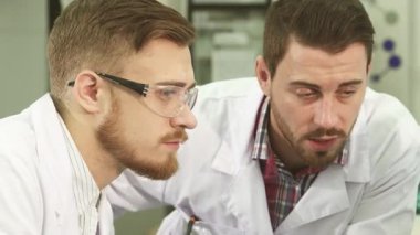 Yakın plan çekimde iki genç erkek laboratuvar teknisyeni ciddi bir sohbete girişiyor, odaklanmış ifadeleri tartışmalarının ciddiyetini ortaya koyuyor. Yüksek teknolojili ekipmanlarla çevrelenmiş, kritik verileri analiz ediyorlar,