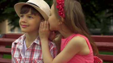 Çekici küçük bir kız, canlı bir park bankında gülümseyen bir çocuğun kulağına hoş bir sır fısıldamak için eğiliyor. Neşeli ifadeleri ve şakacı etkileşimleri çocukluk arkadaşlığının özünü yansıtıyor. Yeşillik ve sıcacık bir ortam ile çevrili.