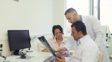 Parlak bir doktor muayenehanesinde genç bir kadın, ciddi bir ifadeyle iki doktor gibi dikkatlice oturur ve röntgen sonuçlarını inceler. Atmosfer profesyonellik ve özen ile dolu.