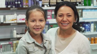 Neşeli bir anne ve genç kızı son zamanlardaki eczane alışverişlerini göstermek için kameraya ışıldıyorlar. Anne şık bir kahverengi çanta tutarken kız gururla bir kredi kartı sergiliyor, neşe ve beraberliği somutlaştırıyor.
