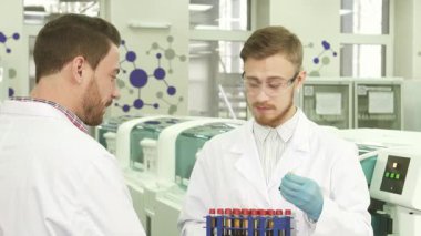 Parlak, modern bir laboratuvarda, iki genç erkek meslektaşı son bulguları hakkında canlı bir tartışmaya giriştiler. Biri laboratuvar önlüğü ve lateks eldiven giyerken diğeri hareketli hareketlerle renkli test tüpleriyle dolu bir standı tutuyor.