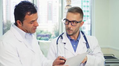 Birinde steteskop, diğerinde de pano olan iki özel doktor, iyi aydınlatılmış bir doktorun muayenehanesindeki hasta sertifikasını dikkatle incelerler. Ciddi ifadeleri, tartışmalarının önemini yansıtıyor.