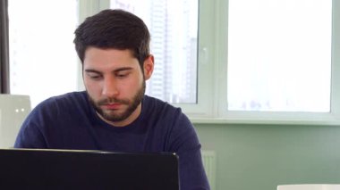 Yakışıklı sakallı bir mimarın yakın plan fotoğrafı. Önünde bir dizüstü bilgisayarla masasında dinlenmek için biraz zaman ayırıyor. Genç beyaz erkek tasarımcı koltuğunda rahatça uzanıyor, profesyonellik ve yaratıcılığın bir karışımını parlak bir şekilde somutlaştırıyor.