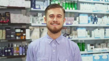 Şık sakallı ve sarı saçlı neşeli genç bir adam parlak bir eczanede başparmağını sallayarak iki alışveriş torbasını gururla sergiliyor. Neşeli ifadesi, sağlık ve güzellik ürünleriyle dolu raflarla çevrili memnuniyet saçıyor.