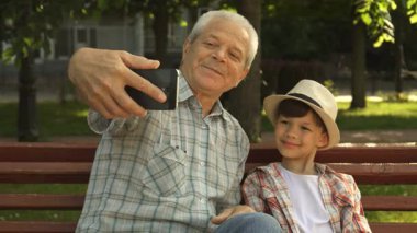 Çekici bir son sınıf öğrencisi, her ikisi de canlı bir park bankında oturan sevimli torunuyla neşeyle selfie çeker. Şık bir şapka takan neşeli küçük çocuk, yemyeşil çimenler ve ağaçlarla çevrili güneşli bir günün tadını çıkarırken mutlulukla parıldıyor.