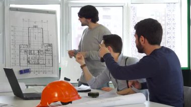 Üç beyaz erkek mimar dinamik bir tartışmaya giriştiler ve bir tablonun ayrıntılı planına işaret ettiler. Sahne, takım çalışmasının özünü yakalıyor, şık bir esmer mühendis, iki sakallı meslektaşından fikir almak istiyor, hepsi de dalmış.