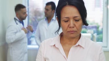 Parlak bir ofiste, elleri şakaklarına bastırılmış genç bir kadın keskin bir baş ağrısından rahatsız olur. İfadesi derin bir rahatsızlığı yansıtırken, beyaz önlüklü iki erkek doktor arka planda sohbet ederek,