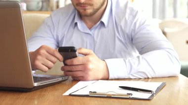 Yakışıklı, sarışın bir işadamı, akıllı telefonuyla derinden meşgul, tahta bir masada oturuyor, pano ve kalemle süslenmiş. Kafenin sıcak atmosferi, verimliliğin rahatlıkla buluştuğu modern bir çalışma alanını gözler önüne sererek sahneyi güzelleştiriyor.