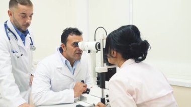 Parlak, modern bir ofiste, iki neşeli doktor bir hastayla ilgilenir, biri gözlerini incelemek için özel bir cihaz kullanır. Ortam sıcak ve davetkar. Dostça sohbetler ve profesyonel ilgiyle dolu.