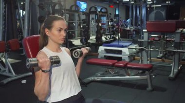 Atkuyruklu, uzun sarı saçlı odaklanmış genç bir kadın dambılları kaldırıyor, gücünü ve bağlılığını gösteriyor. Spor malzemeleriyle çevrili, her temsilcisiyle limitlerini zorlayan fitness ve kararlılık ruhunu somutlaştırıyor.