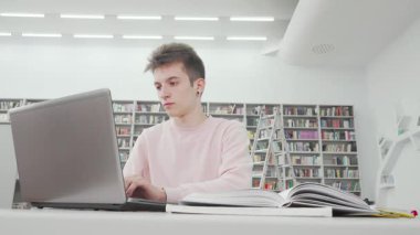 Düşük açılı bir çekim, kendini işine adamış bir erkek öğrenciyi parlak ve çağdaş bir kütüphanede dizüstü bilgisayarıyla derinden bağlı bir şekilde yakalar. Yüksek kitap raflarıyla çevrelenmiş, akademik arayış ruhunu somutlaştırıyor, teknoloji ve eğitimin füzyonunu gösteriyor.