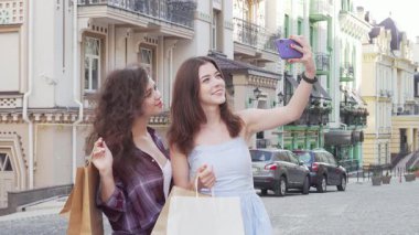 İki güzel bayan arkadaş, ellerinde çantalar, güzel bir şehir zemininde akıllı telefonlarıyla selfie çekiyorlar. Gülüşleri ve enerjik enerjileri dostluk ve macera saçıyor. Bu anı tasasız bir anın mükemmel bir görüntüsü yapıyor.