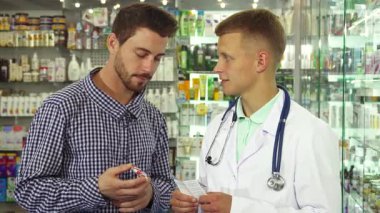 Beyaz önlüklü bilgili bir doktor, rehberlik isterken elinde bir şişe hap tutan meraklı bir erkek hastayla karşılaşır. Renkli ilaç ürünlerinin raflarıyla çevrelenmiş bu sahne, sağlık hizmetlerinin ve hastanın özünü yakalıyor.