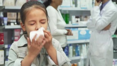 Hareketli bir eczanede, yorgun küçük bir kız mendile hapşırır, yorgun gözleri rahatsızlığını yansıtır. Arkasında, annesi renkli sağlık ürünleri raflarıyla çevrili bir eczacıyla sohbet ediyor.