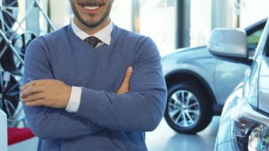 İyi bakımlı sakallı ve koyu renk saçlı neşeli bir adam kendine güvenerek duruyor, arabanın anahtarlarını göğsüne yakın tutuyor. Bir elini kaldırırken gülümsemesi heyecan saçıyor. Yeni arabasının anahtarlarını sergiliyor. Modern bir galeride gösterişli araçlarla çevrili.