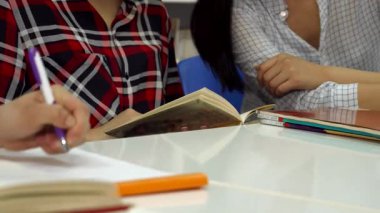 Yakın plan çekimde, derslerine dalmış öğrencilerle çevrili bir erkek eli, bir deftere zarifçe yazarken görülüyor. Sahne, bir kadın eliyle zarifçe bir kitabın sayfasını çeviriyor ve canlı bir öğrenme atmosferi yaratıyor.