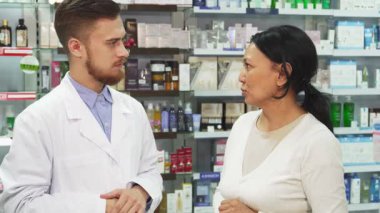 Parlak bir eczanede, neşeli bir kadın elinde bir alışveriş çantasıyla durup, alışverişini ilgili genç bir eczacıdan memnuniyetle alır. Her ikisi de dostça bir sohbete girişip sıcak bir gülümseme sergiliyor.
