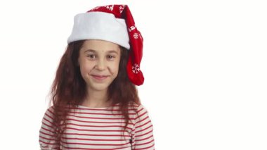 Neşeli kız kameraya ışıldıyor, kıvırcık siyah saçları sevinçle dönerken zıplıyor. Klasik bir Noel Baba şapkası takıp beyaz bir arka plana yaslanarak bayram havası ve heyecan saçar.