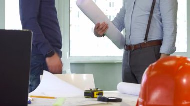 Gösterişli bir ofis ortamında, açık mavi gömlekli ve pantolon askılı bir erkek mimar iş arkadaşına bir tomar bina planı verir. Sahne, takım çalışmasının ve yaratıcılığın özünü, sert bir şapka ve çizim araçlarıyla yakalıyor.