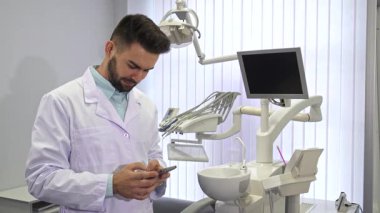 Yakışıklı genç bir dişçi, düşünceli bir ifadeyle, modern bir diş muayenehanesinde dururken akıllı telefonunda bir numara çeviriyor. Gıcır gıcır beyaz önlük giyen şık esmer erkek doktor gelişmiş diş ekipmanlarıyla çevrili.