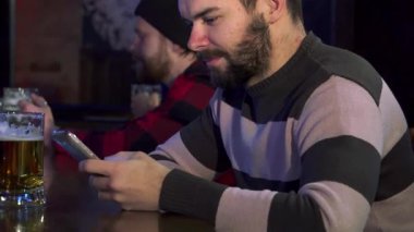 Şık sakallı çekici bir genç adam, sıcak ortam aydınlatmasıyla aydınlatılmış, hareketli bir barda akıllı telefonunu çeviriyor. Sahne, modern sosyal etkileşimin özünü yakalıyor, sosyal medyada gezinirken aynı zamanda
