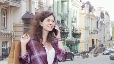 Hoş, genç bir kadın şehir caddesinde canlı bir telefon görüşmesi yapıyor, alışveriş çantaları yanında sallanıyor. Işıl ışıl bir gülümseme ve şık bir kıyafetle şehir keşfi ve perakende terapisinin neşesini somutlaştırıyor.