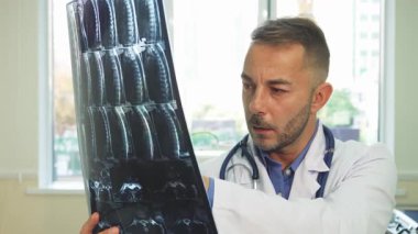 Gıcır gıcır beyaz bir ceket giymiş genç bir erkek doktor parlak ofisinde röntgen filmini inceledikten sonra kameraya ışınlanıyor. Sıcak atmosfer, geniş pencerelerden süzülen doğal ışıkla güçlenir. Hasta bakımına olan bağlılığını yansıtır.