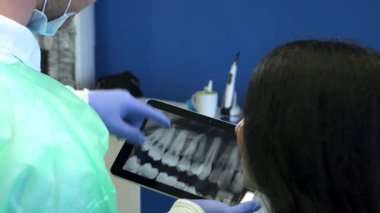 Yakın plan çekimde yeşil üniformalı yakışıklı bir dişçi, beyaz bir kadına tablet üzerinde detaylı bir röntgen sunarken görülüyor. Kıdemli esmer kadın yüzünü doktora doğru çeviriyor, parlak bir nişanda bir anlık özen gösteriyor.